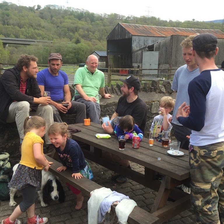 A family sit around a wooden picnic table with drinks on it, large rusty metal farm sheds and woodland in the background.