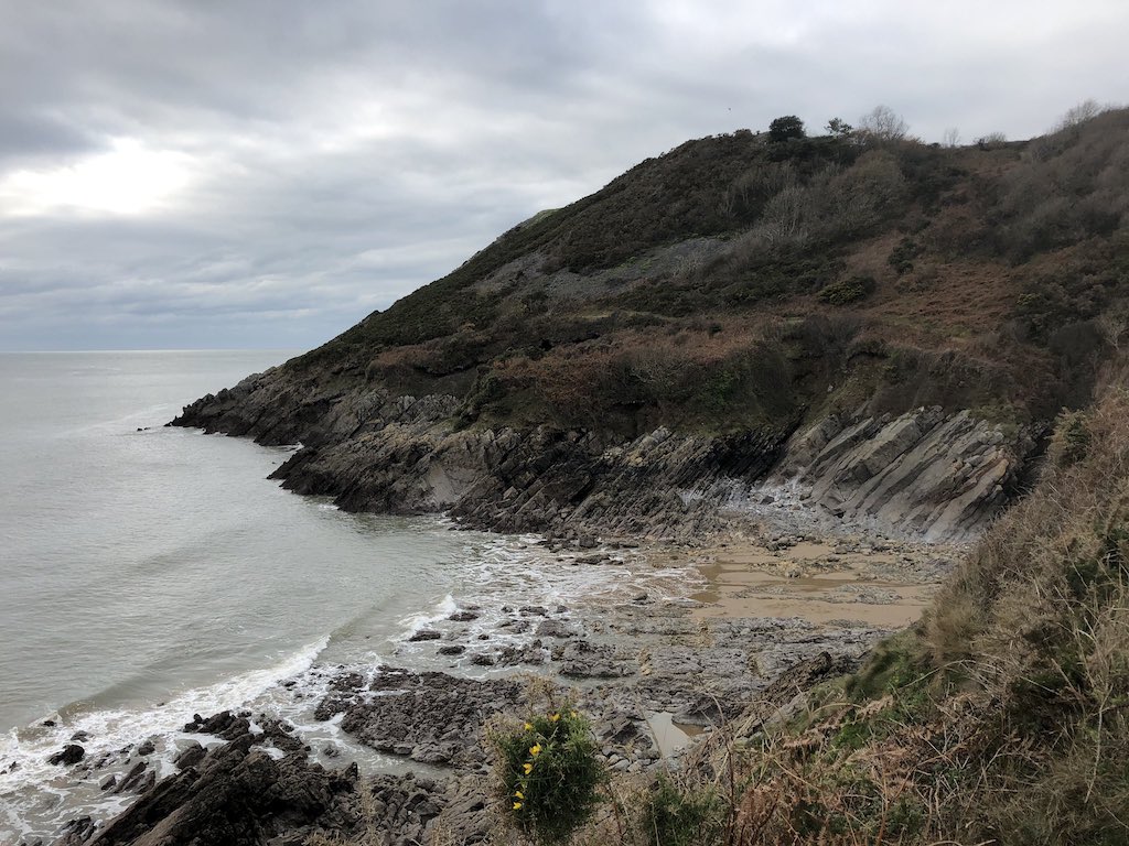 View down from a coastal path to a small sandy and rocky beach. Small waves break from the left. Cliffs on the far side lead up to dark green hills with shrubs and bushes.