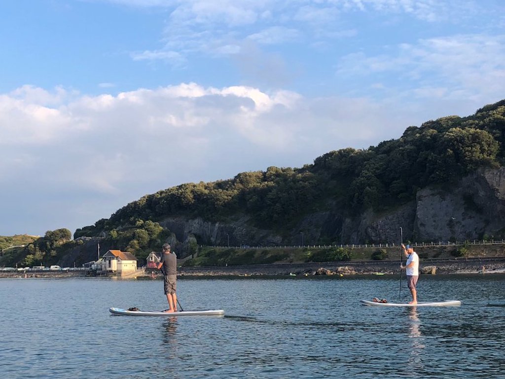 Two paddle boarders on the sea. Cliffs topped with trees in the background. Sky is cloudy and blue.
