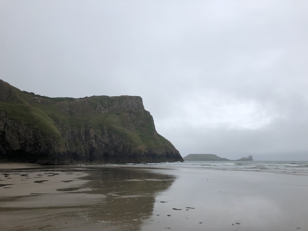 Steep cliffs topped with green grass reflect in wet sand below. Sky is grey.