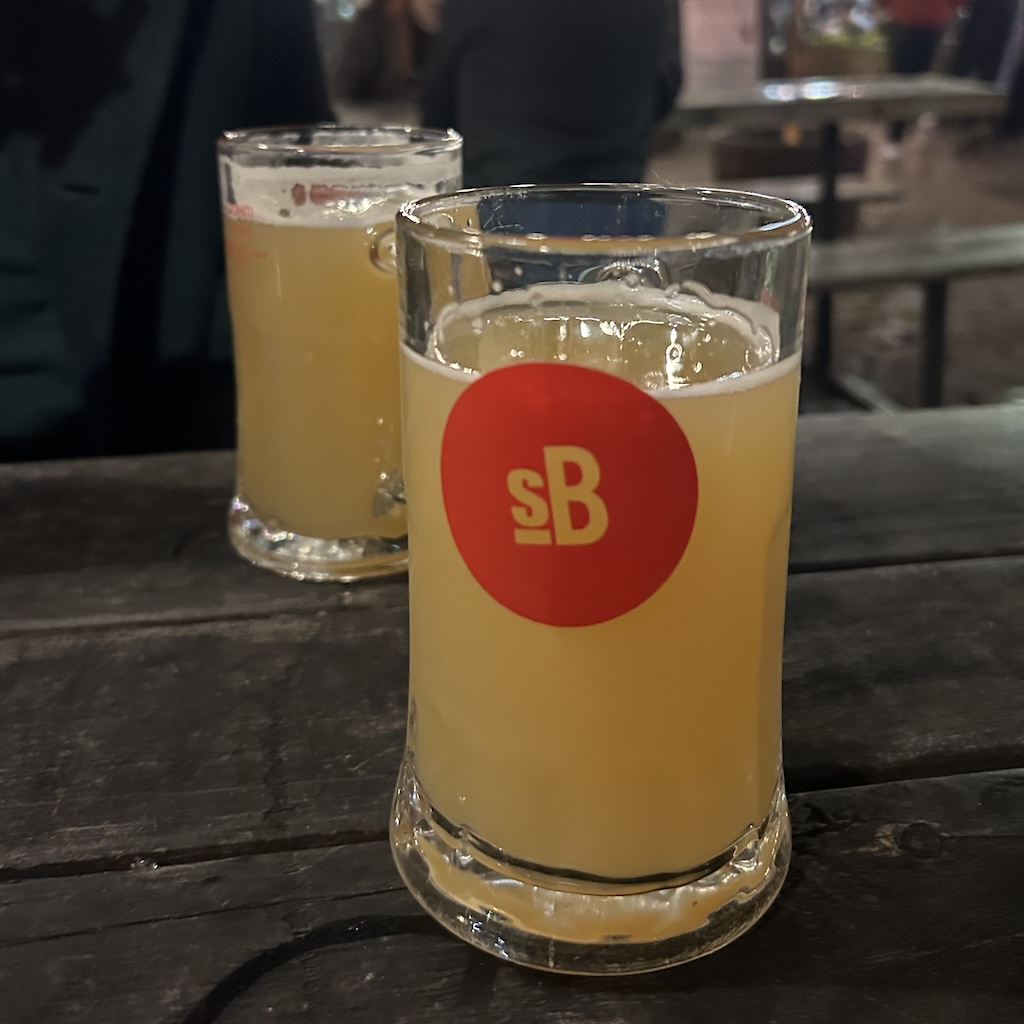 A couple of two-third size glasses filled with hazy yellow beer. One has a red circle printed on it with the letters s and b inside it.