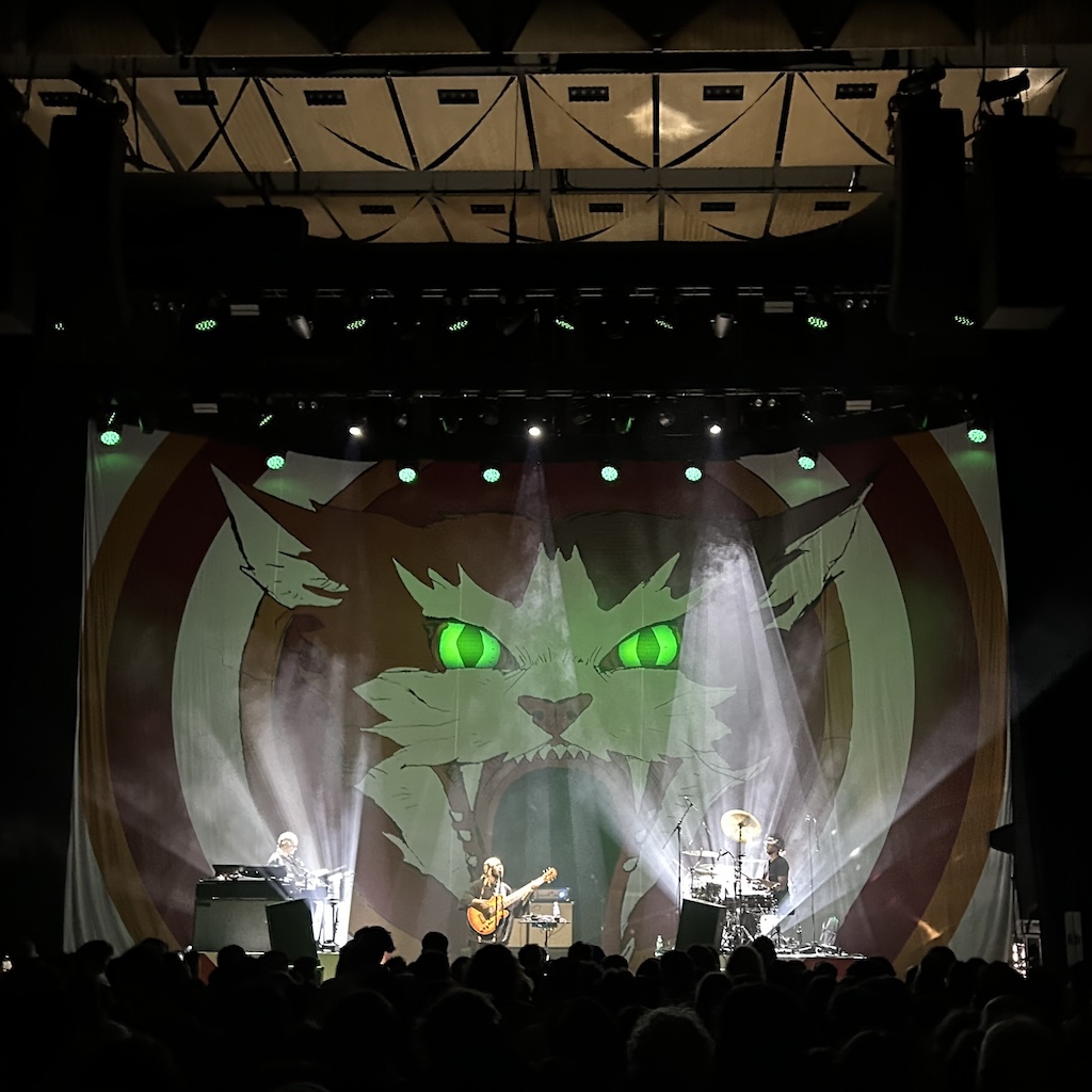 3 people on a stage. A huge, fierce cat face in the background has glowing green eyes.