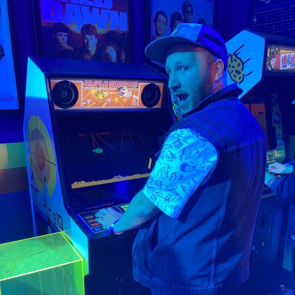 Me in a dark room, tinged in blue light, playing an 80s style missile command arcade machine. I’m turning round to look at the camera. Wearing a cut off black denim jacket and a baseball cap.
