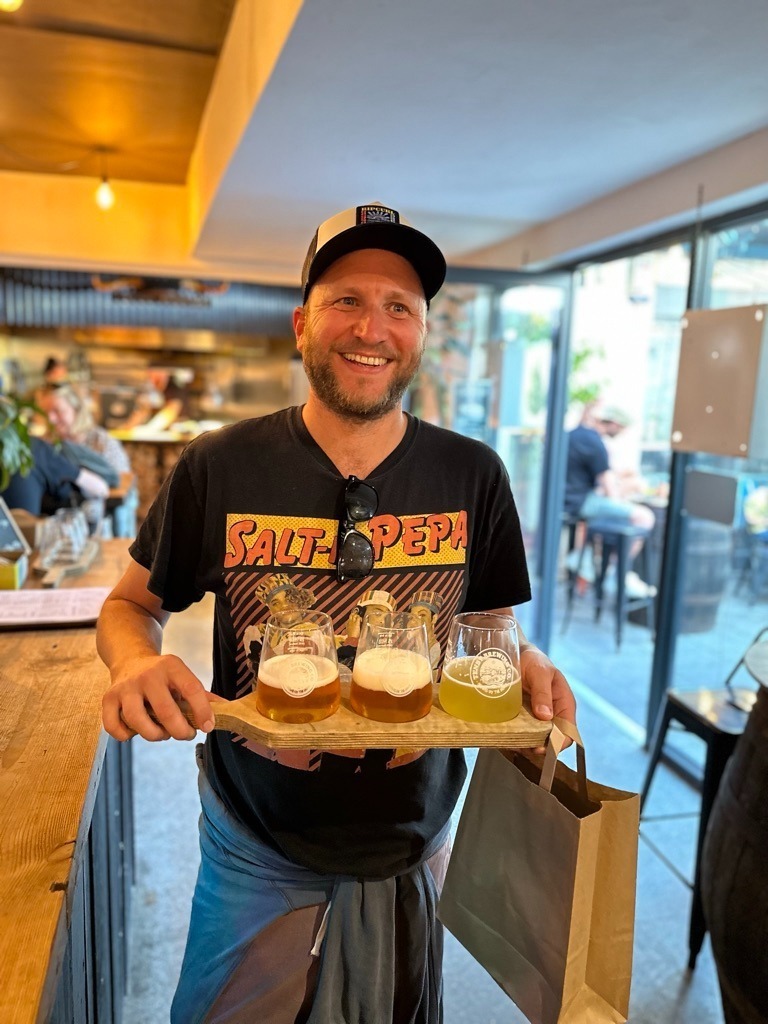 White man with beard and cap, smiling and looking very happy, carrying a wooden flight with 3 beers on it