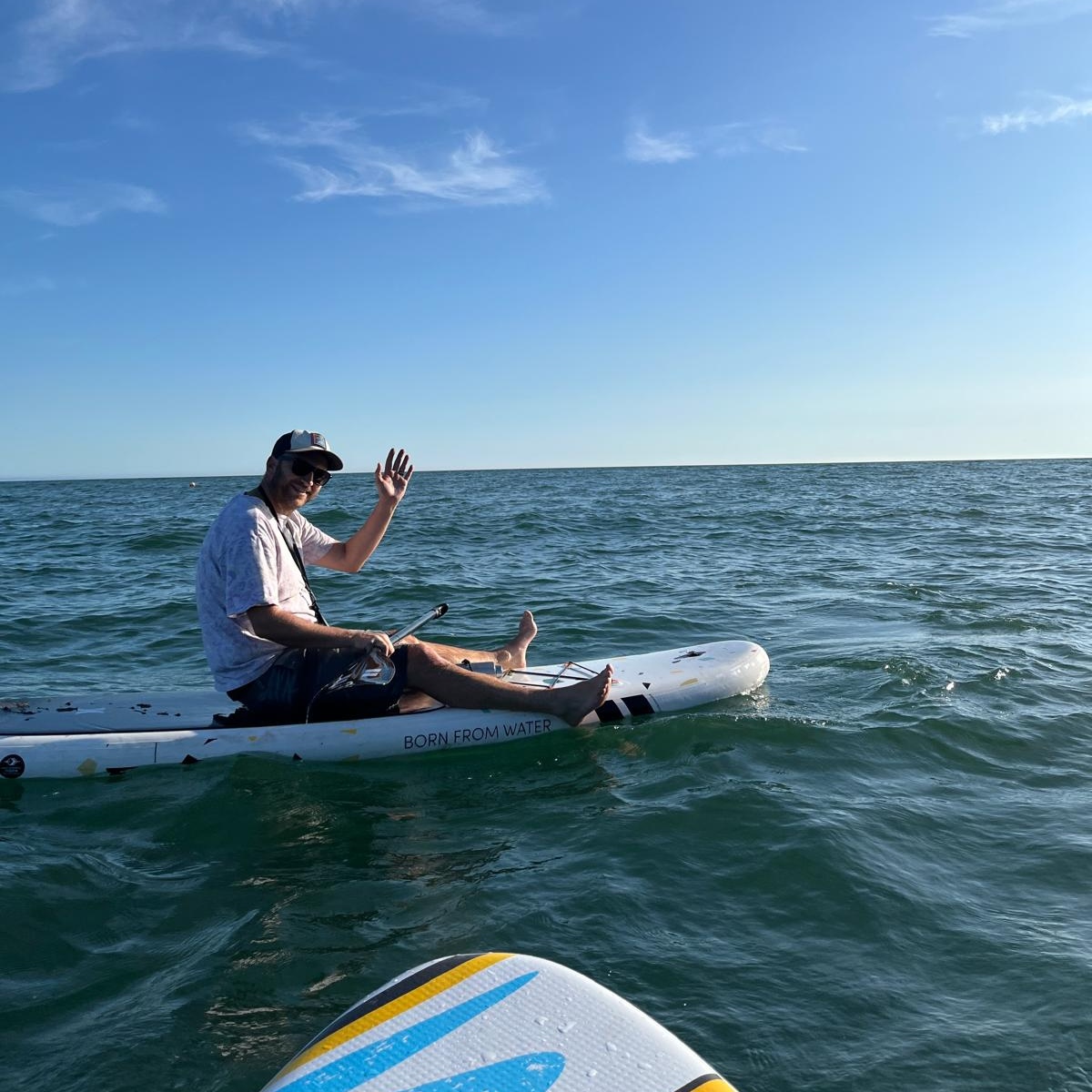 A man sits on a paddle board, waving at the camera. There is only sea and sky in the background.