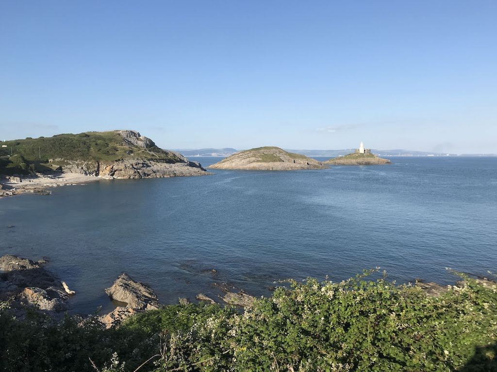 A rocky bay at high tide. 3 mounds of headland stretch into the sea. The last has a white lighthouse on top. The sky is blue and clear.