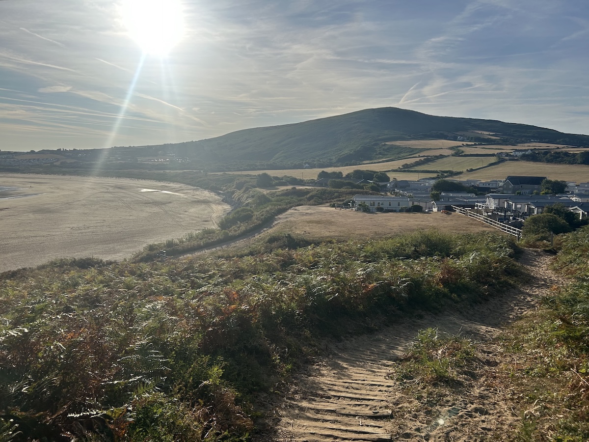 A beach side path through ferns towards a caravan site. Rolling fields and hills in the distance.