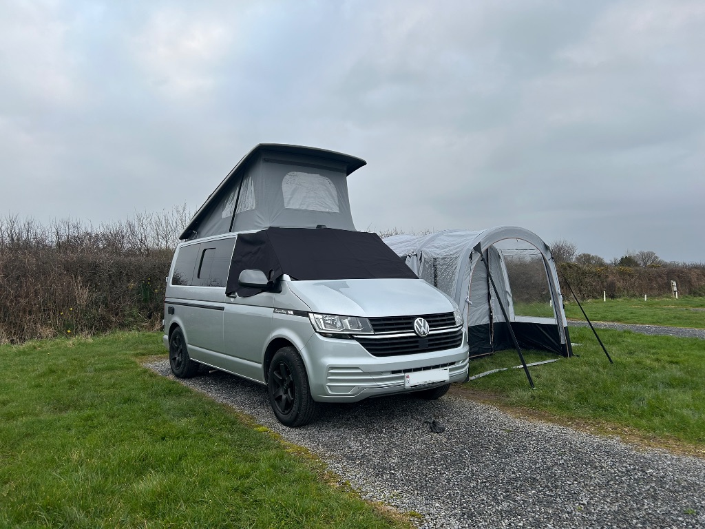 A silver camper van in a grassy field with the roof up and an awning attached. A small black dog poo bag sits on the gravel pitch at the front.