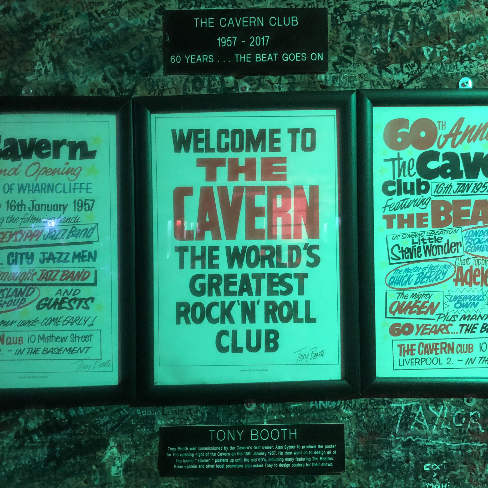 Framed posters on a wall. The middle one says: welcome to the cavern the world’s greatest rock ‘n’ roll club. A small plaque below it says they were designed by Tony Booth. The brick wall in the background is completely covered in signatures. The room is quite dark but bathed in cool, blue light.