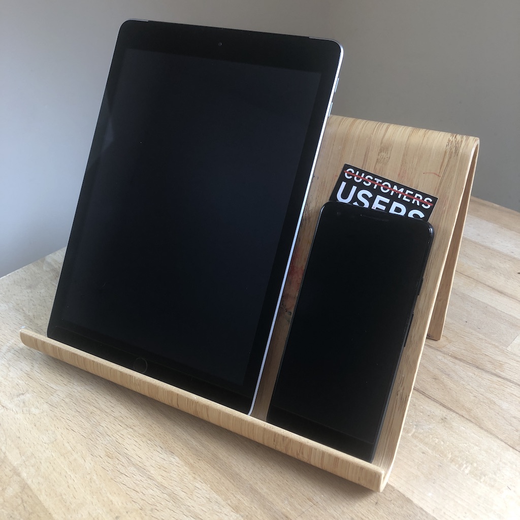 An iPad and a Google Pixel phone rest on a wooden stand on a table. Behind the phone, a black sticker with white text that says "Customers, Users" The word customer has a red line through it.