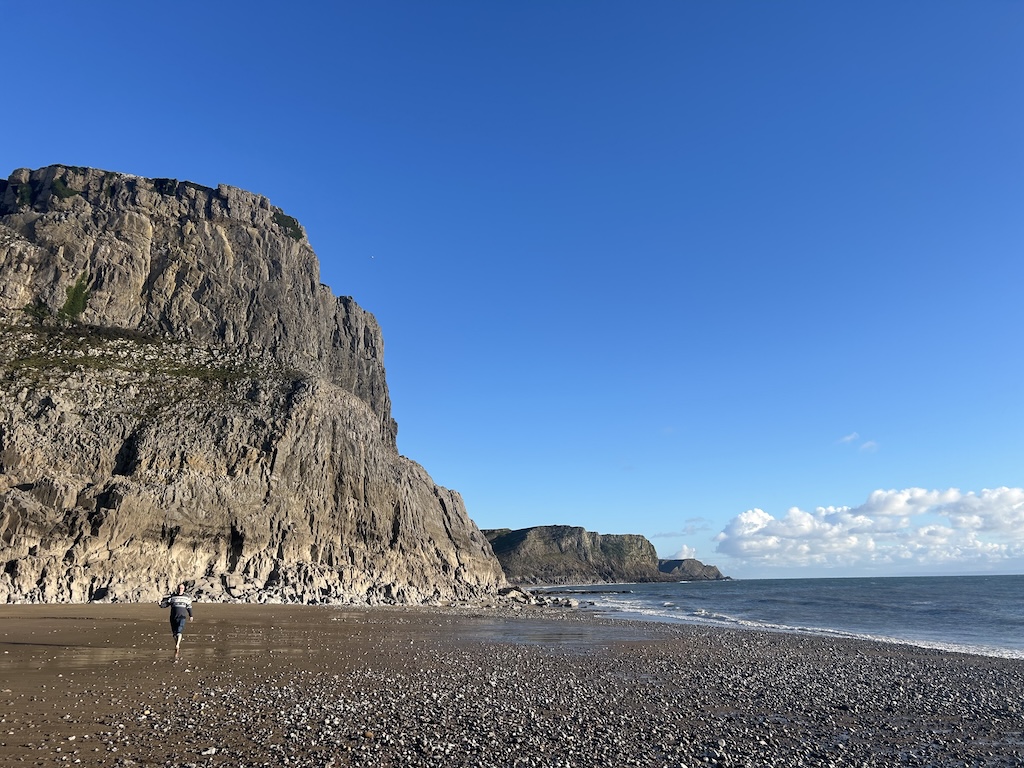 Pebbly beach with tall, steep rocky cliffs. A man walks in the distance. The sky is very blue with a few clouds.