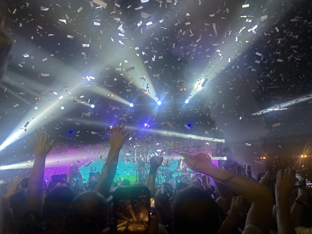 A crowd watching a gig. Hands are waving in the air as bright lights and confetti fill the room.