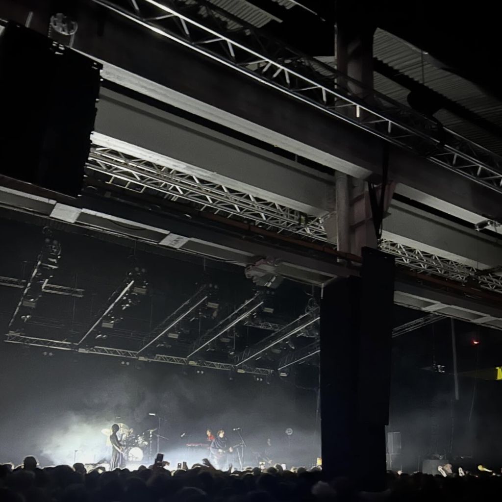Band playing on a stage in a huge warehouse. They are backlit by bright white lights and smoke.