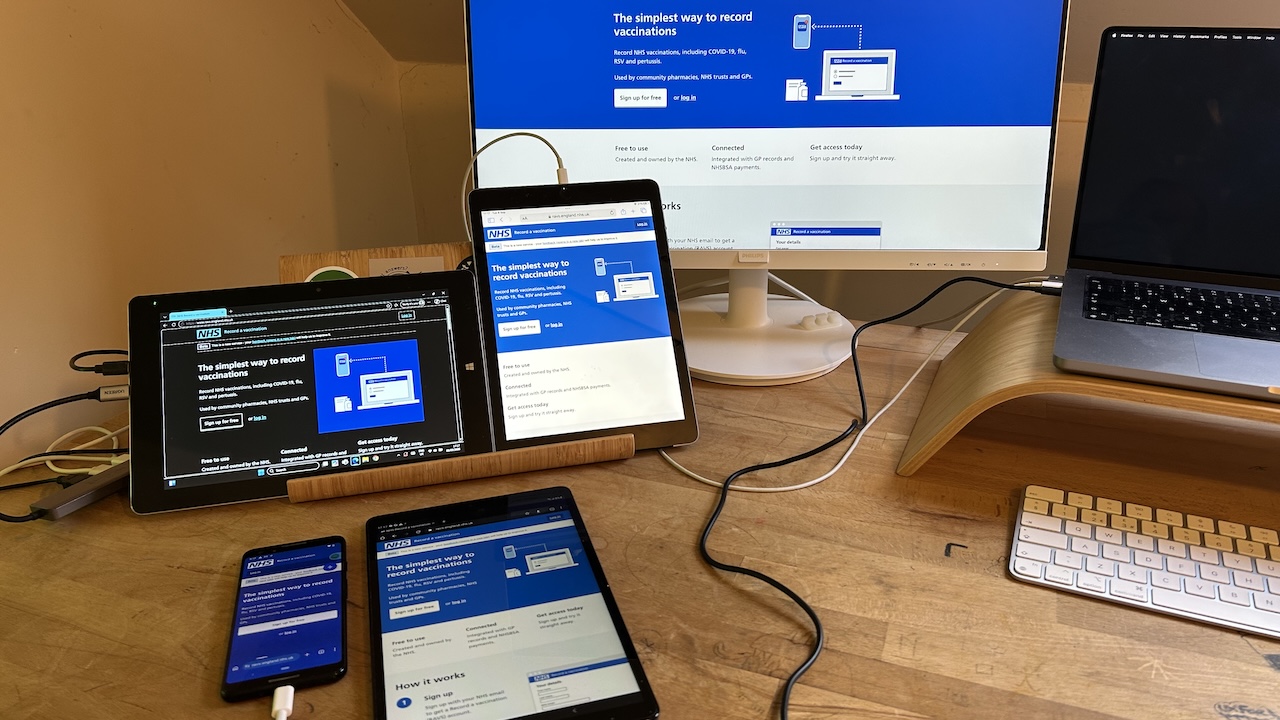 A wooden table with a desktop display, various tablets and a phone all showing the same website.