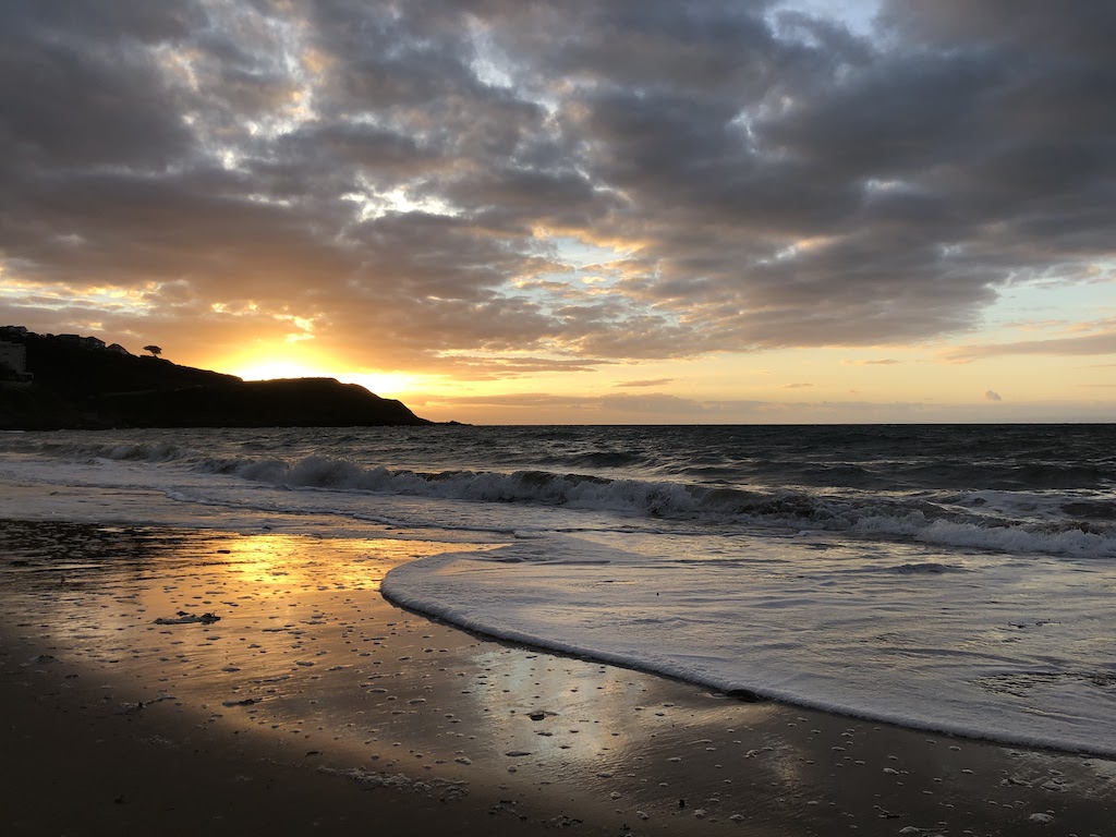 Waves on a sandy shoreline. Sunrise behind the headland in the distance is reflecting orange in the wet sand.