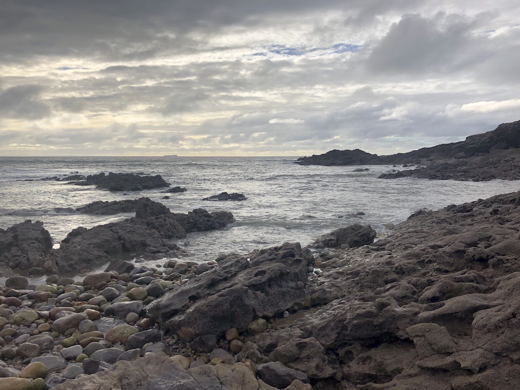 Grey rocks and pebbles disappear into the sea at the shore. Jagged rocks poke out of the sea, and rocky headland can be seen in the distance.