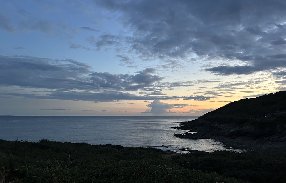A small cove at dusk. The land is dark but the sea and sky are still bright. The horizon has some orange tones from the setting sun which is out of sight.