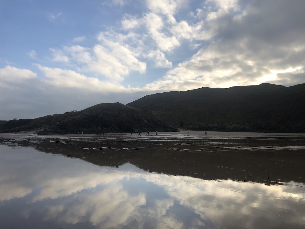 A massive stretch of wet sand with grass topped cliffs in the distance. The cliffs and sky are mirrored almost perfectly in the wet sand below.