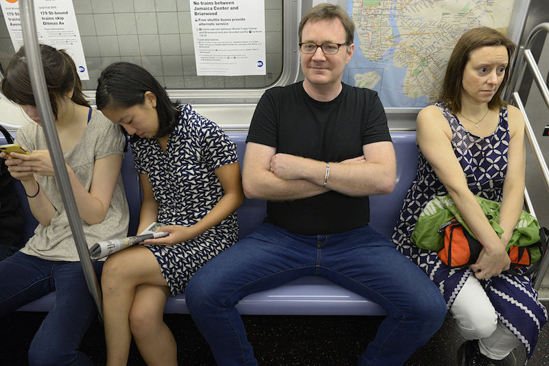 Man on a train sitting with his legs very far apart.