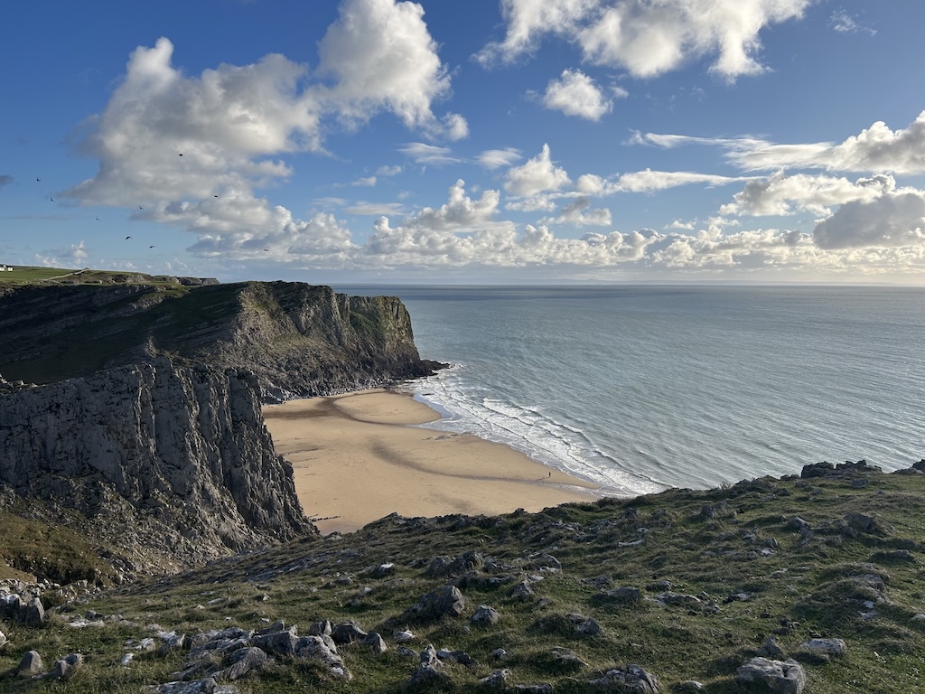 Looking down towards a sandy beach at low tide from high cliffs. The cliffs extend into the sea at the far side.
