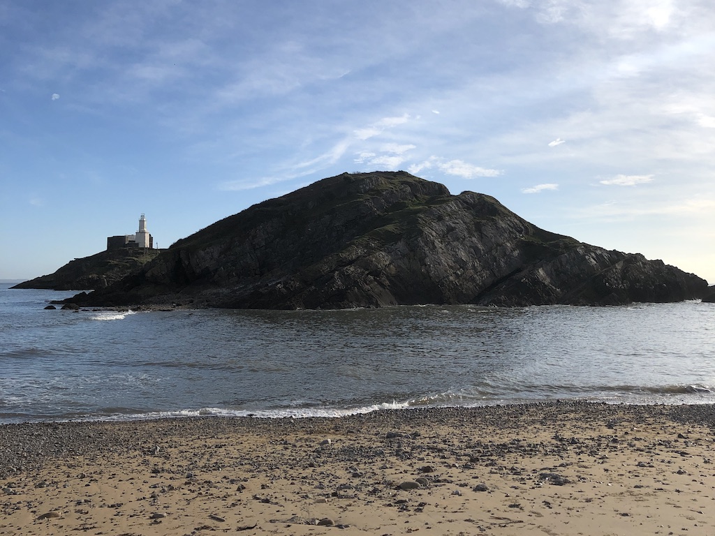 Small rocky island with a lighthouse, just the other side of the water from a sandy beach.