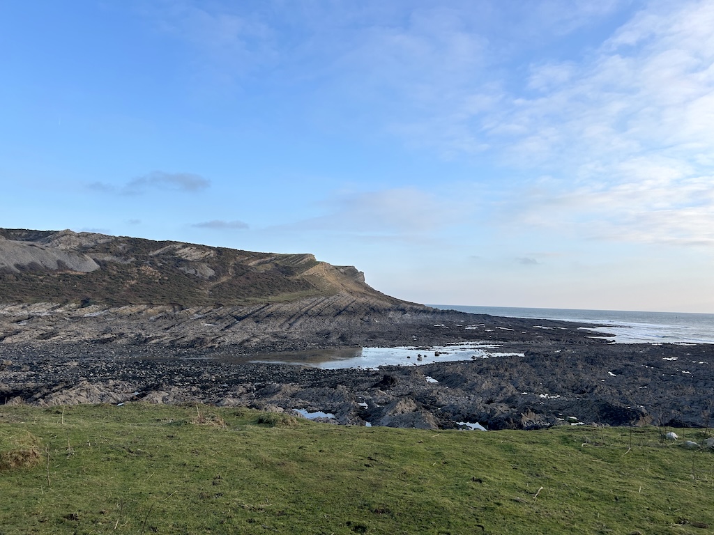 A grassy ledge in the foreground. A large rocky cove with huge rock pools. Green cliffs in the background and blue sky.