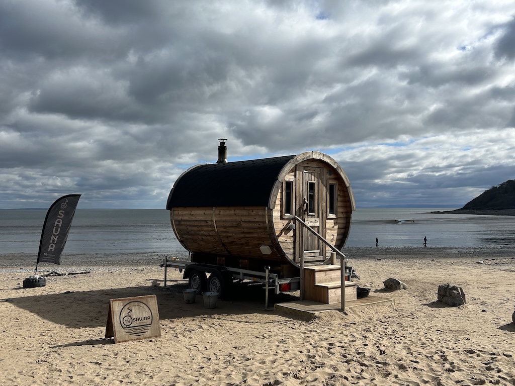 A wooden barrel sauna on a trailer on the beach. The tide is out in the background and it's cloudy.