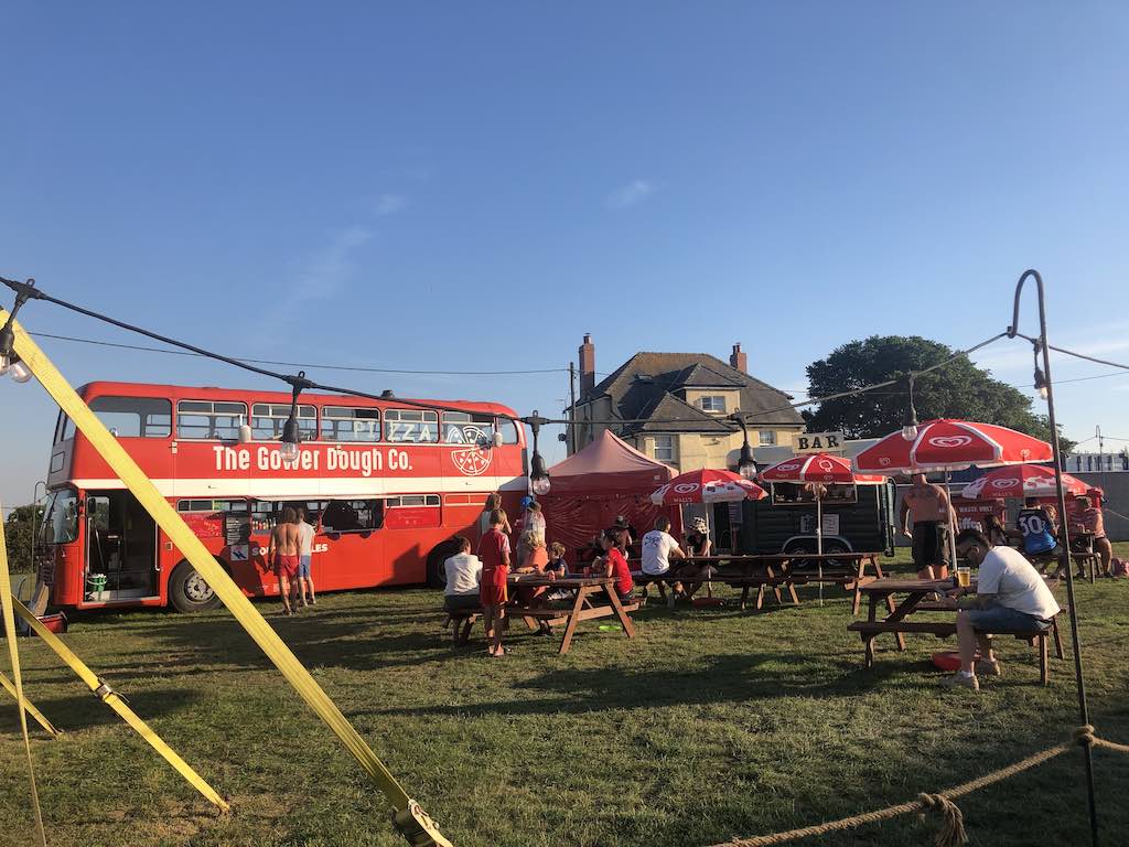 Red double decker bus, serving pizzas in a green field, surrounded by picnic benches with red umbrellas. A large house can be seen in the background.