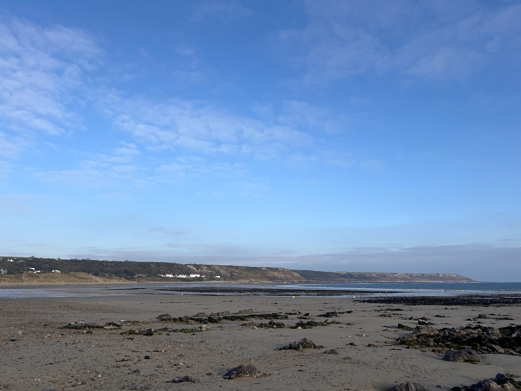 A vast sandy beach at low tide with sand dunes, white houses and green headland in the distance.