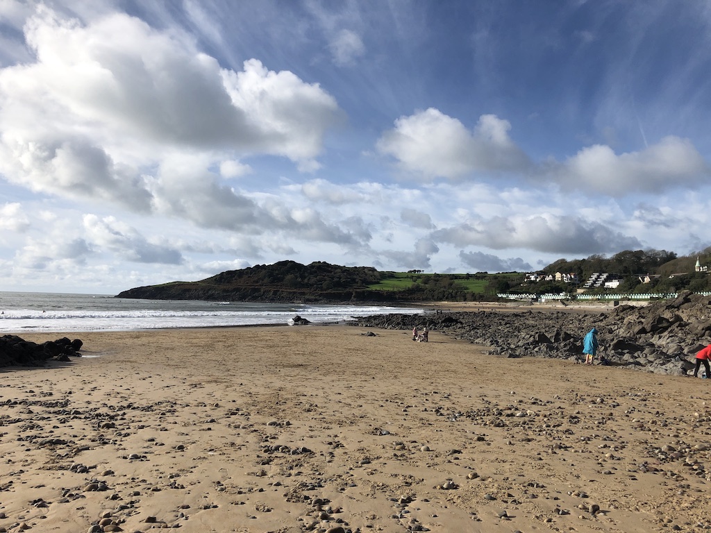 A sandy, pebbly beach at low tide. Beyond the rocks on one side, another beach and green hills can be seen.