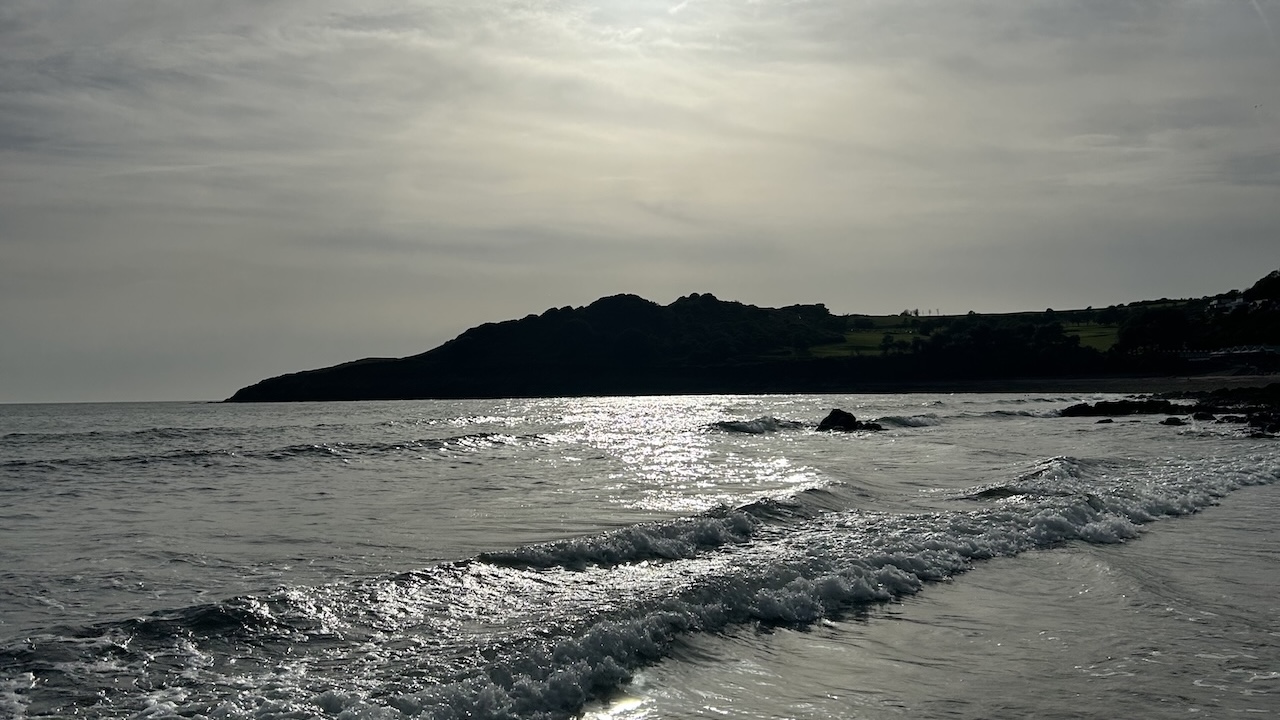A beach in the late afternoon. The sun is low and reflecting in the glassy water. The headland in the distance is almost black against the bright setting sun.