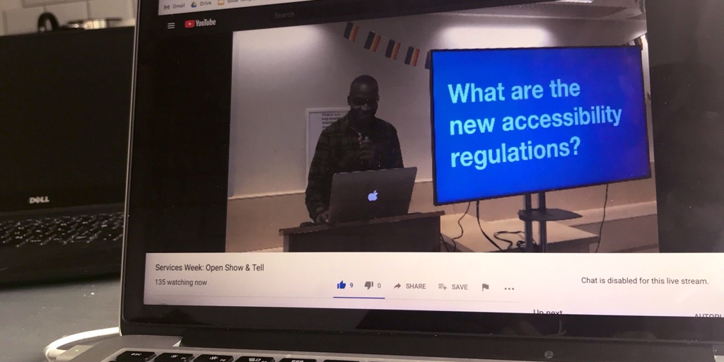 A laptop showing a live stream from London. A designer stands next to a slide that says “what are the new accessibility regulations?”.