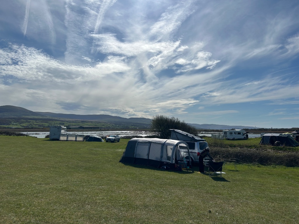 Campervan and awning in a green field with reflective water, rolling hills and blue cloudy sky.