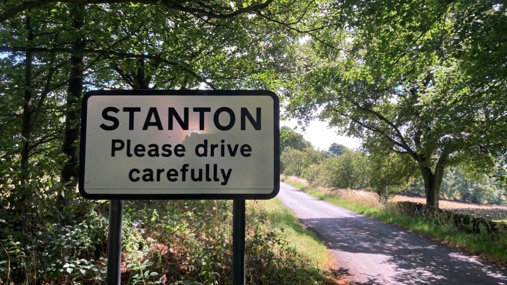 A black and white road sign says: STANTON Please drive carefully. It’s beside a narrow road with trees either side.