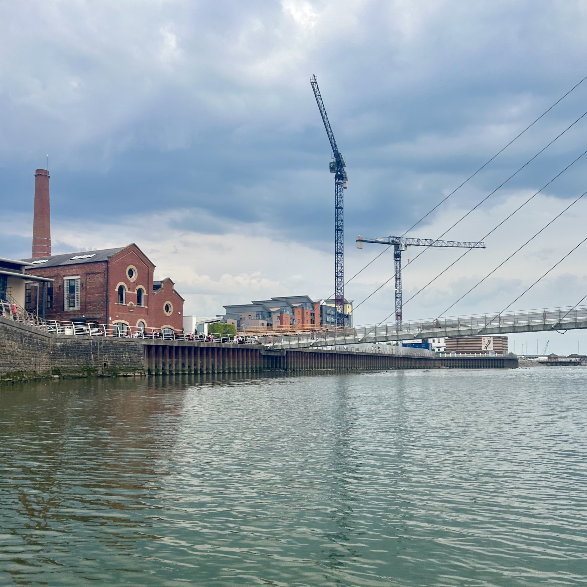 Old red brick buildings and new office blocks, cranes and a foot bridge viewed from the water.