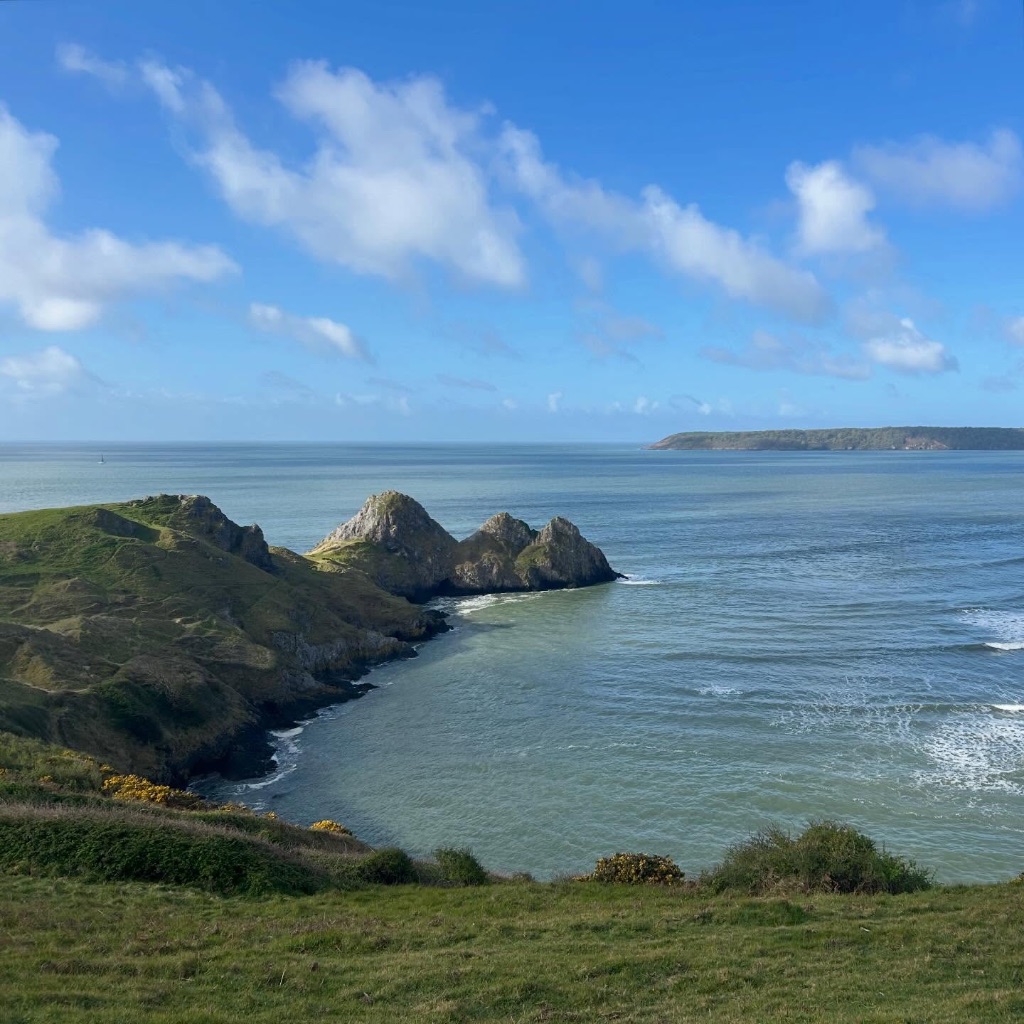 A rocky and grassy headland stretching out into the sea. The tip has 3 peaks. The skyline with wispy clouds.