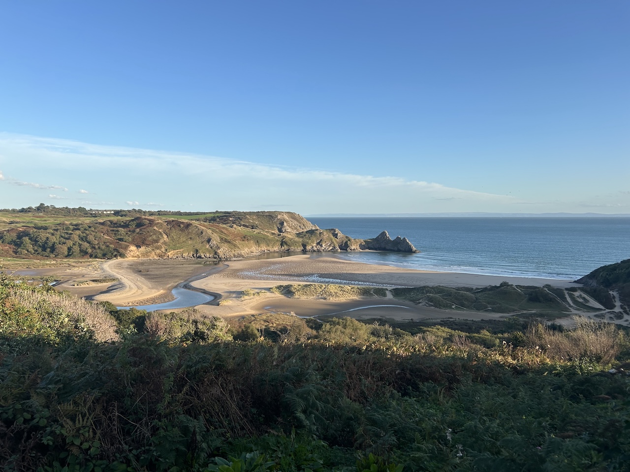 A picturesque sandy beach with a river running through the middle. 3 peaked cliffs reach into the sea.