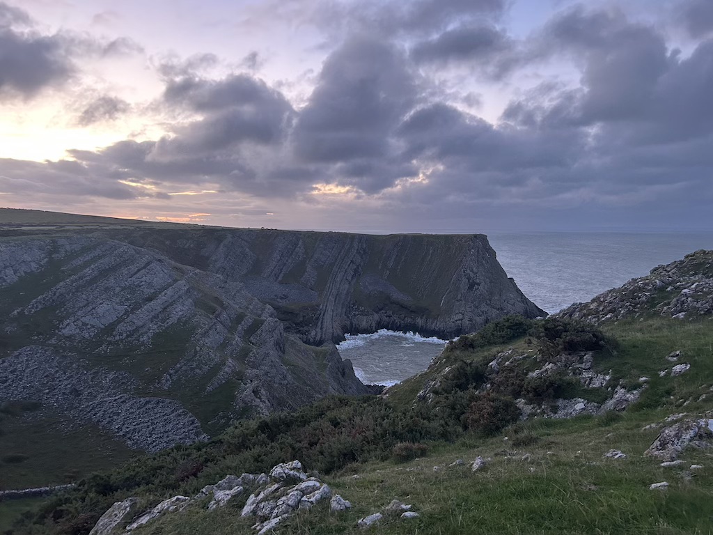 Looking down into a rocky bay with no sand. It's surrounded by tall cliffs. The rocks are almost purple in the earlu morning light.