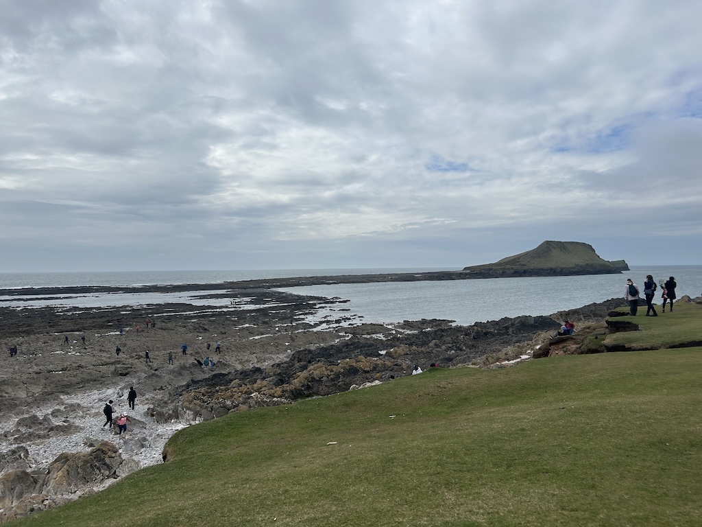 People crossing a rocky causeway across the sea, towards a large grassy peninsula in the distance.